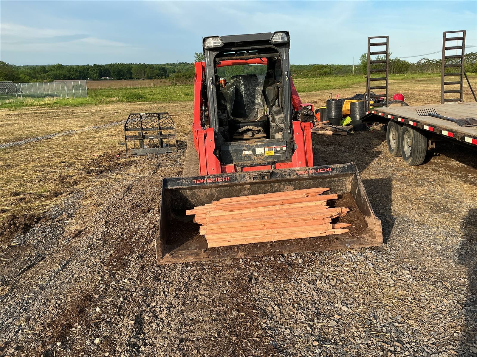 Skid steer carrying planting stakes on a gravel staging area beside a flatbed trailer